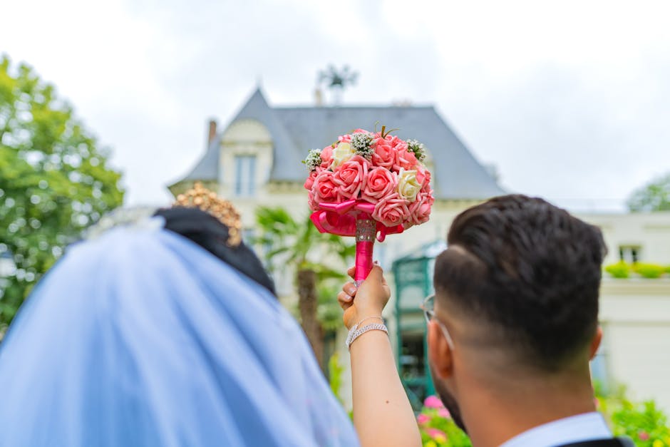 A close-up view of a floral bouquet featuring pink and white roses arranged in a rounded, compact style, with fresh petals and green foliage. The bouquet is wrapped with a bright pink ribbon and held