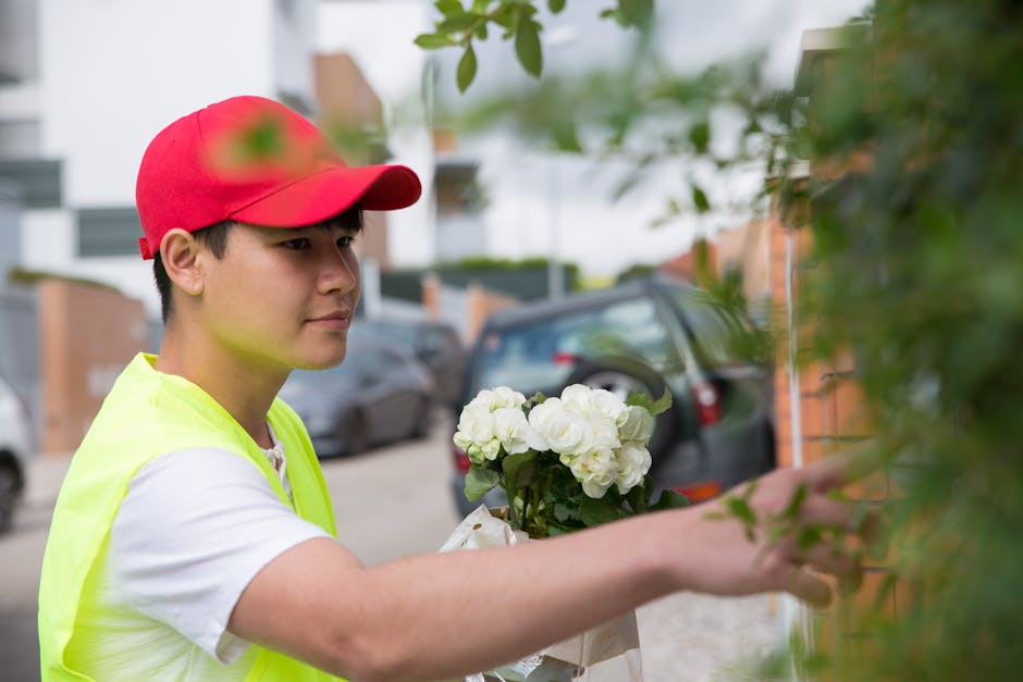 A young male florist with light skin, wearing a bright red cap and a high-visibility yellow vest over a white t-shirt, arranging a bouquet of white hydrangeas outdoors. He is reaching out to place or 